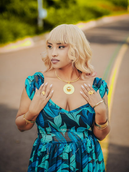 A woman with blonde hair and bangs wearing a blue and green tropical print Habesha chiffion, gold jewelry, and a blonde wig, posing outdoors on a paved road with green foliage in the background.