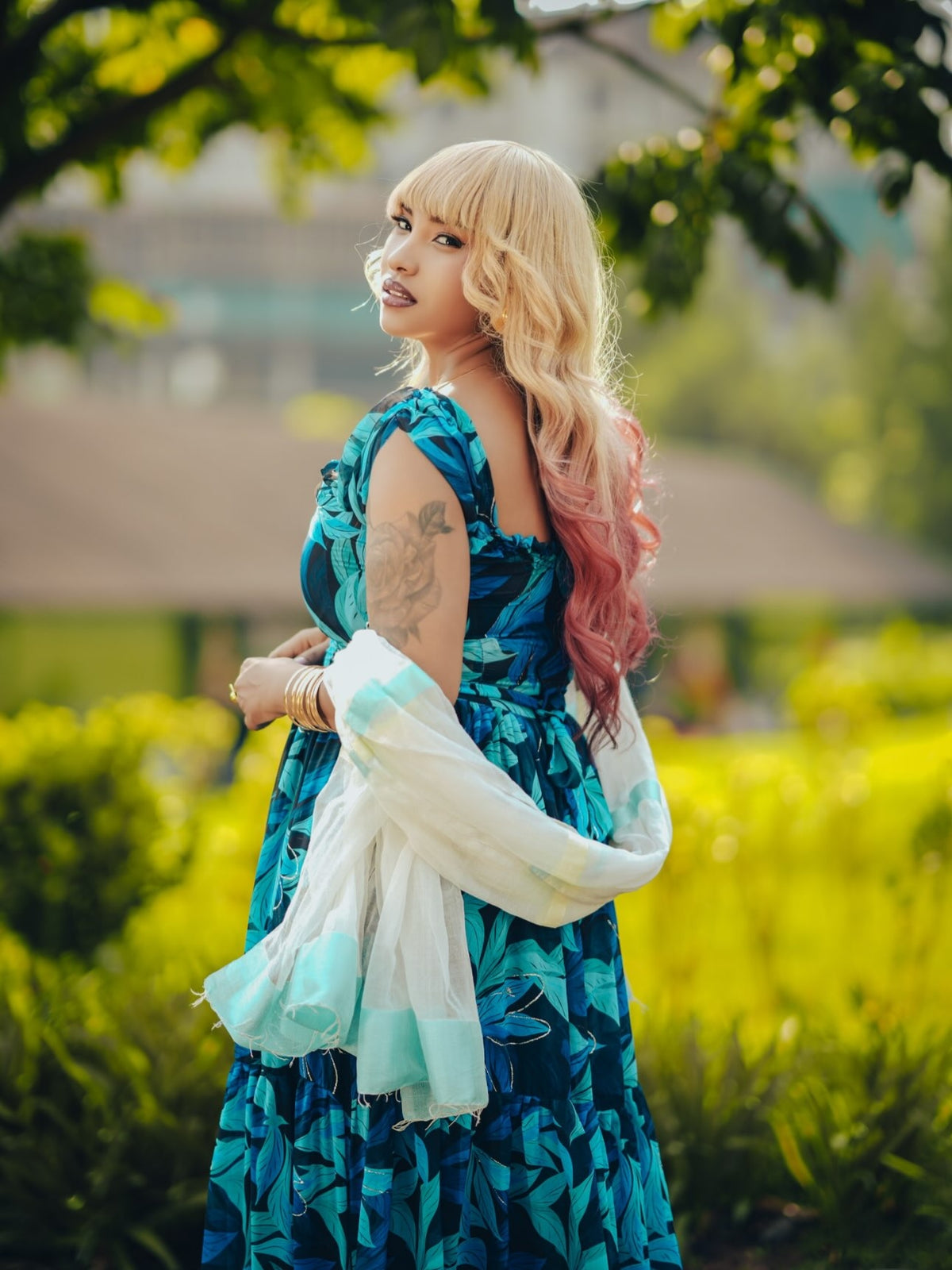 A woman with blonde and pink ombre hair stands outdoors in a park wearing a blue patterned maxi Habesha chiffion with a white sheer scarf draped over her arm.