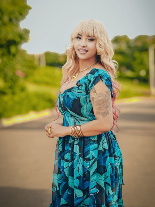 A woman with blonde hair and pink tips stands outdoors wearing a blue and teal leaf-patterned habesha chiffion, gold jewelry, and a gold necklace. The background shows a blurred green park setting.