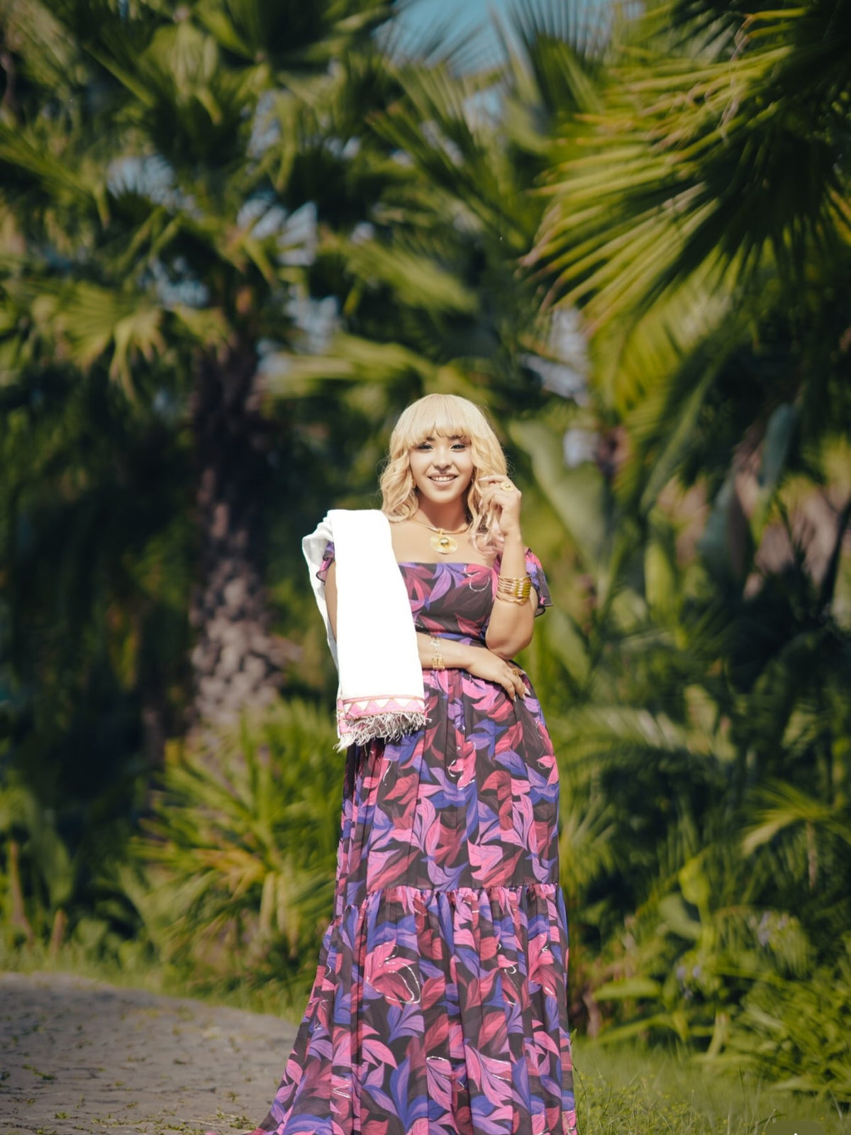 A woman stands outdoors in a tropical garden with palm trees, wearing a long sleeveless habesha chiffon with a pink, purple, and blue floral pattern. She is holding a white towel draped over her arm.