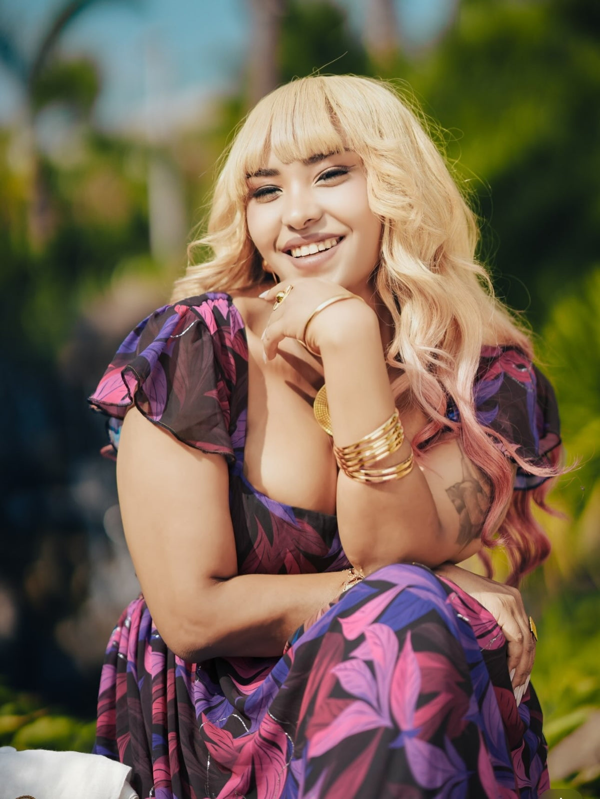 A woman with long blonde hair and bangs sits outdoors wearing a loose, deep V-neck habesha chiffon with a purple, pink, and black floral pattern. She is wearing multiple gold bracelets and has a warm smile on a blurred green background.