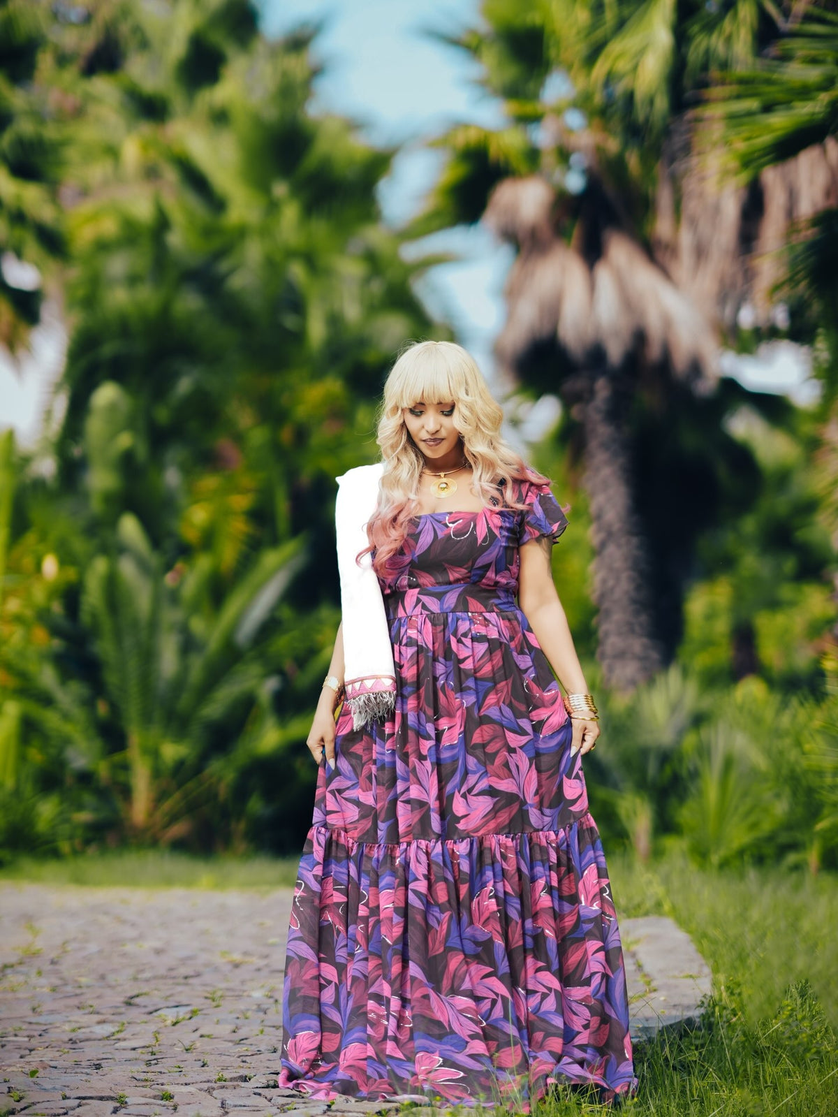 A woman stands outdoors on a stone path wearing a long tiered habesha chiffon with a pink, purple, and blue floral pattern, styled with a white sleeveless cardigan. The background is a lush green tropical garden with palm trees.