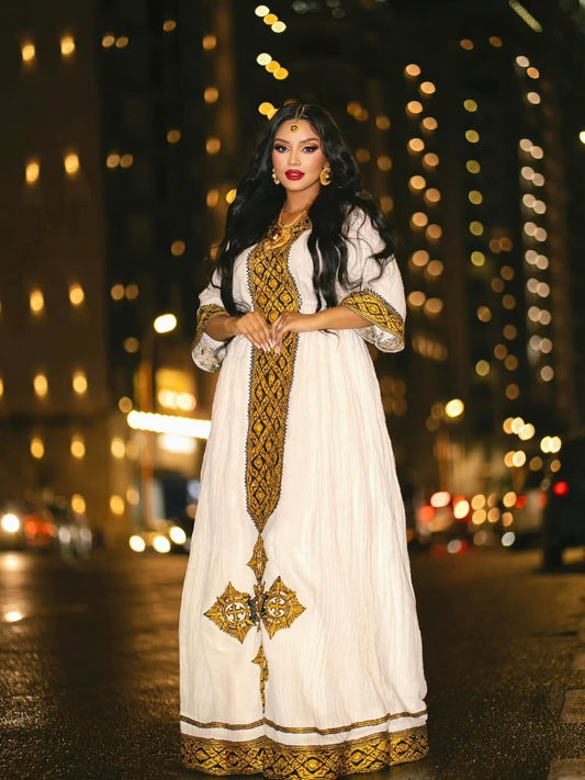 Woman in a white and gold habesha dress standing in a city street at night.