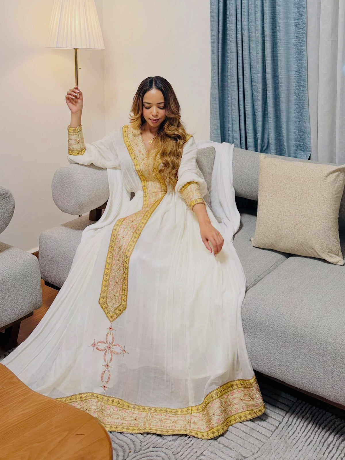 A woman wearing a white traditional Habesha dress  with gold embroidery sits on a gray sofa in a living room setting.