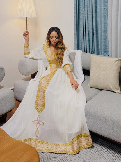 A woman wearing a white traditional Habesha dress  with gold embroidery sits on a gray sofa in a living room setting.