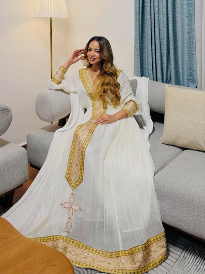 A woman wearing a white traditional Habesha dress  with gold embroidery and a matching dupatta, seated on a gray sofa in a living room setting.