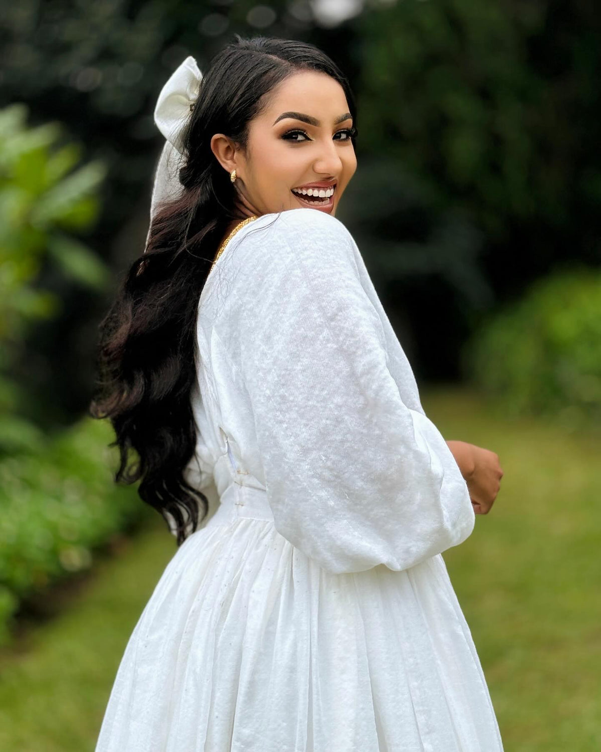 A woman wearing a white long-sleeve textured Ethiopia Store with a matching white bow in her hair, photographed outdoors in a garden setting.