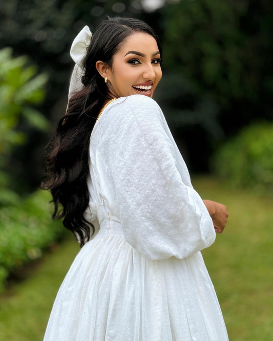 A woman wearing a white long-sleeve textured Ethiopia Store with a matching white bow in her hair, photographed outdoors in a garden setting.
