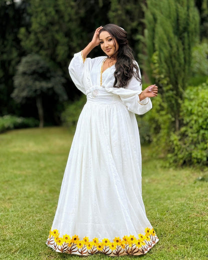 A woman wearing a long white maxi Ethiopia Store with yellow sunflower embroidery along the hem, standing on a green grass lawn with trees in the background.