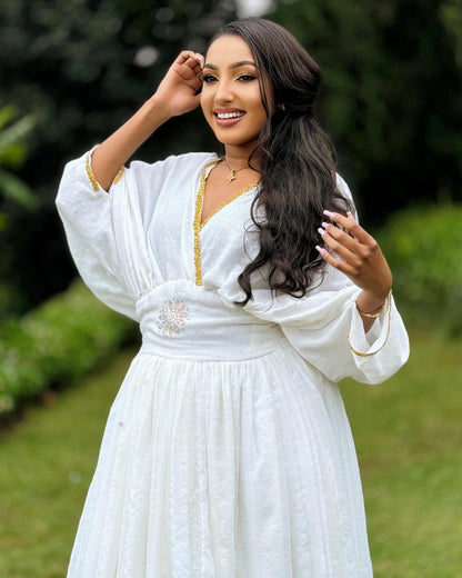 A woman wearing a white long-sleeve Ethiopia Store with gold trim and embroidery, standing outdoors in a garden setting.