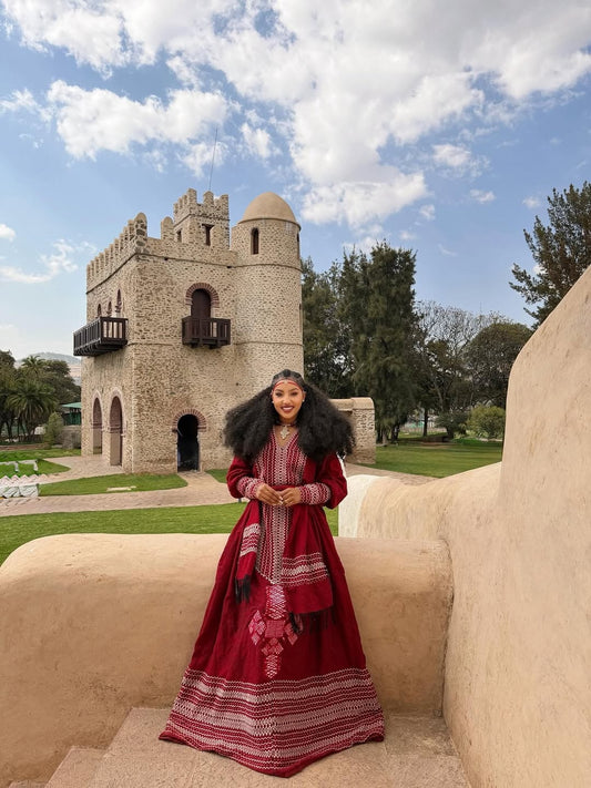 Woman in a red traditional Ethiopian dress standing in front of a stone building with a blue sky.