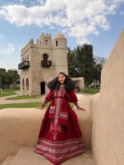 Woman in a red Ethiopian dress standing in front of a stone building with a blue sky.