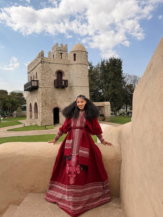 Woman in a red Ethiopian dress standing in front of a stone building with a blue sky.