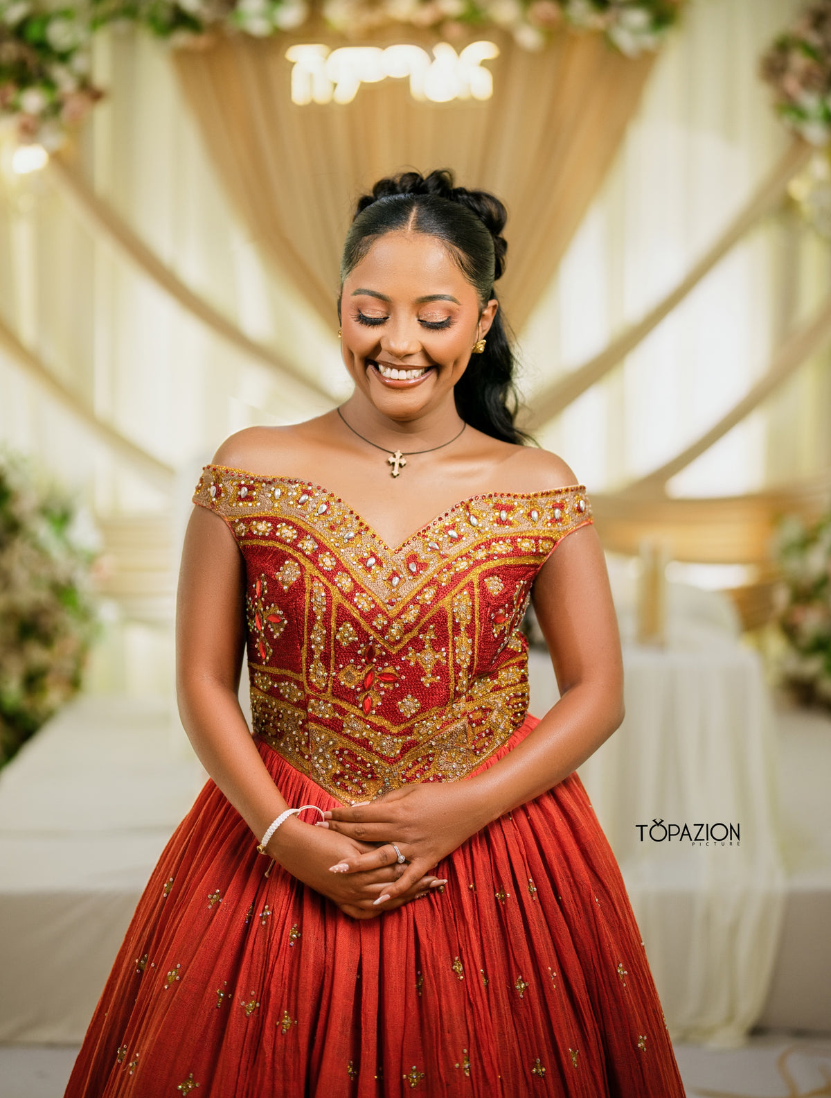 A woman wearing a red off-the-shoulder Habesha Wedding Dress  with extensive gold embroidery and embellishments, standing in front of a decorated cream-colored event backdrop.
