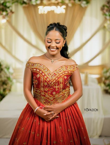 A woman wearing a red off-the-shoulder Habesha Wedding Dress  with extensive gold embroidery and embellishments, standing in front of a decorated cream-colored event backdrop.