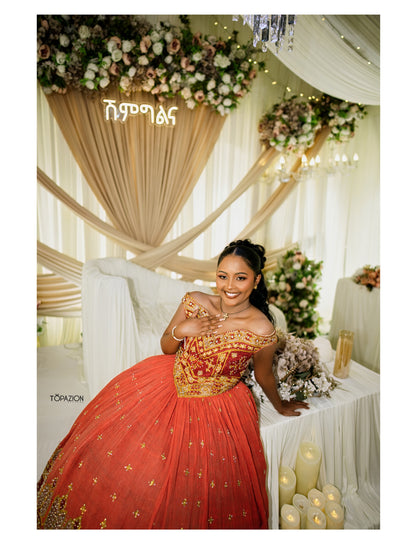 A woman wearing a voluminous off-the-shoulder red and gold traditional ball Habesha Wedding Dress  poses in an elegantly decorated indoor event space with draped cream fabric, floral arrangements, and candles.