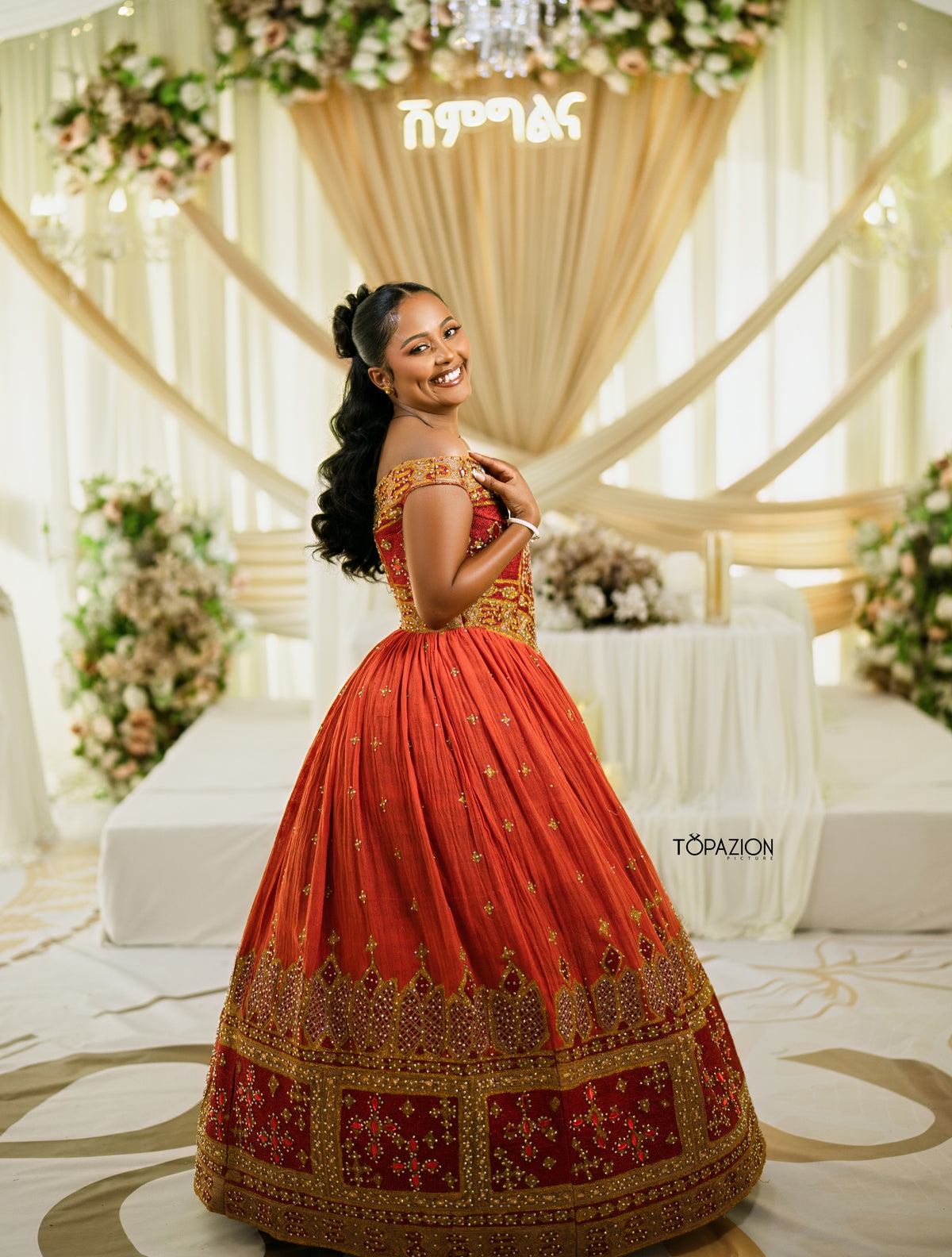 A woman wearing a traditional Indian red and gold lehenga choli Habesha Wedding Dress , posing in a decorated event hall with draped cream fabric and floral arrangements. The brand name 'TOPAZION' is visible as a watermark on the right side.