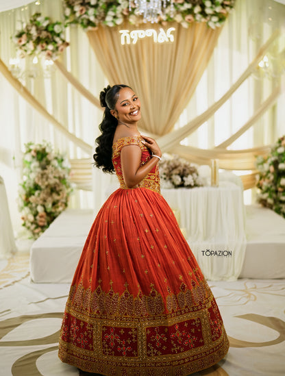 A woman wearing a traditional Indian red and gold lehenga choli Habesha Wedding Dress , posing in a decorated event hall with draped cream fabric and floral arrangements. The brand name 'TOPAZION' is visible as a watermark on the right side.