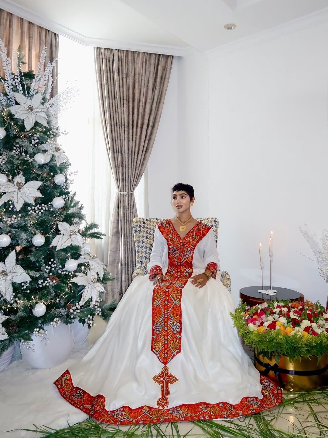 Woman in a traditional Ethiopian traditional dress sitting in a decorated room with a Christmas tree and flowers.