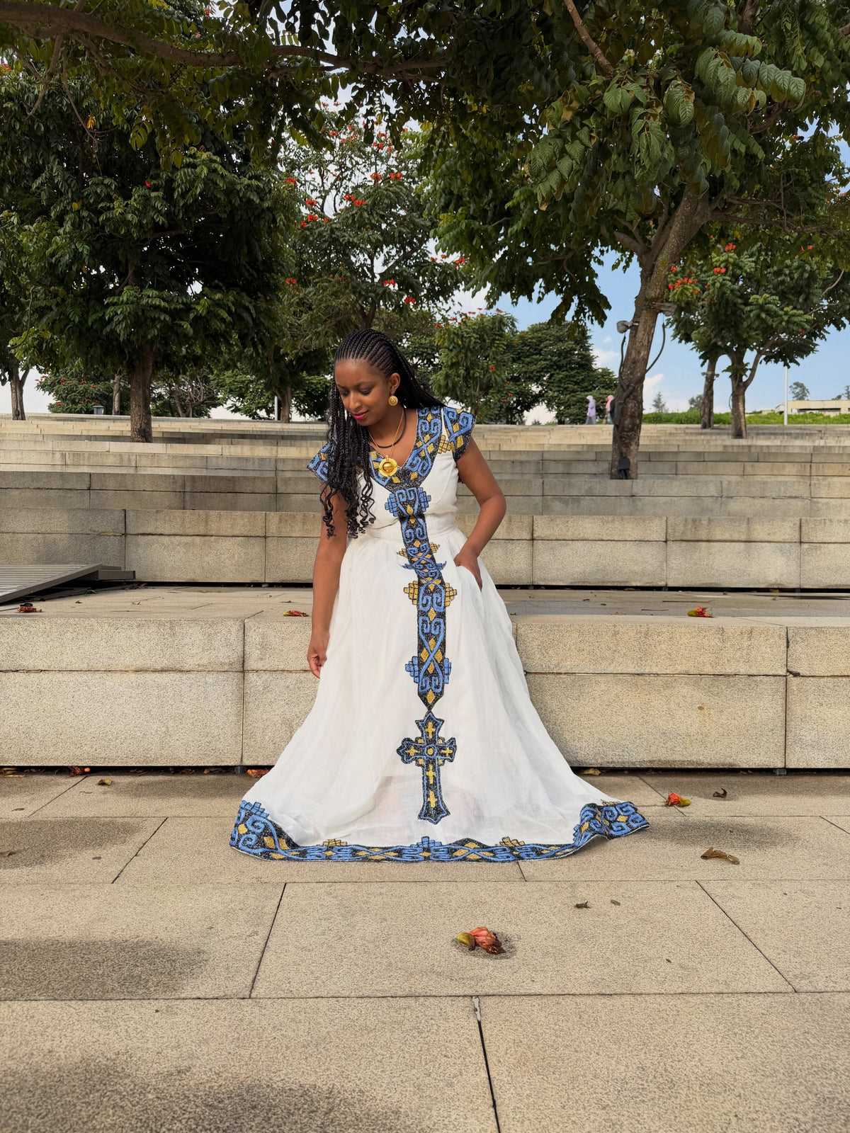 Woman standing outdoors in a white Ethiopian Dress featuring blue and gold woven embroidery down the front center and along the hem.