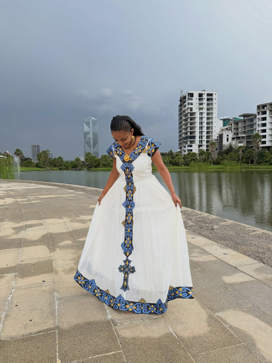 Woman standing outdoors in a white Ethiopian Dress featuring blue and gold woven embroidery down the front center and along the hem.
