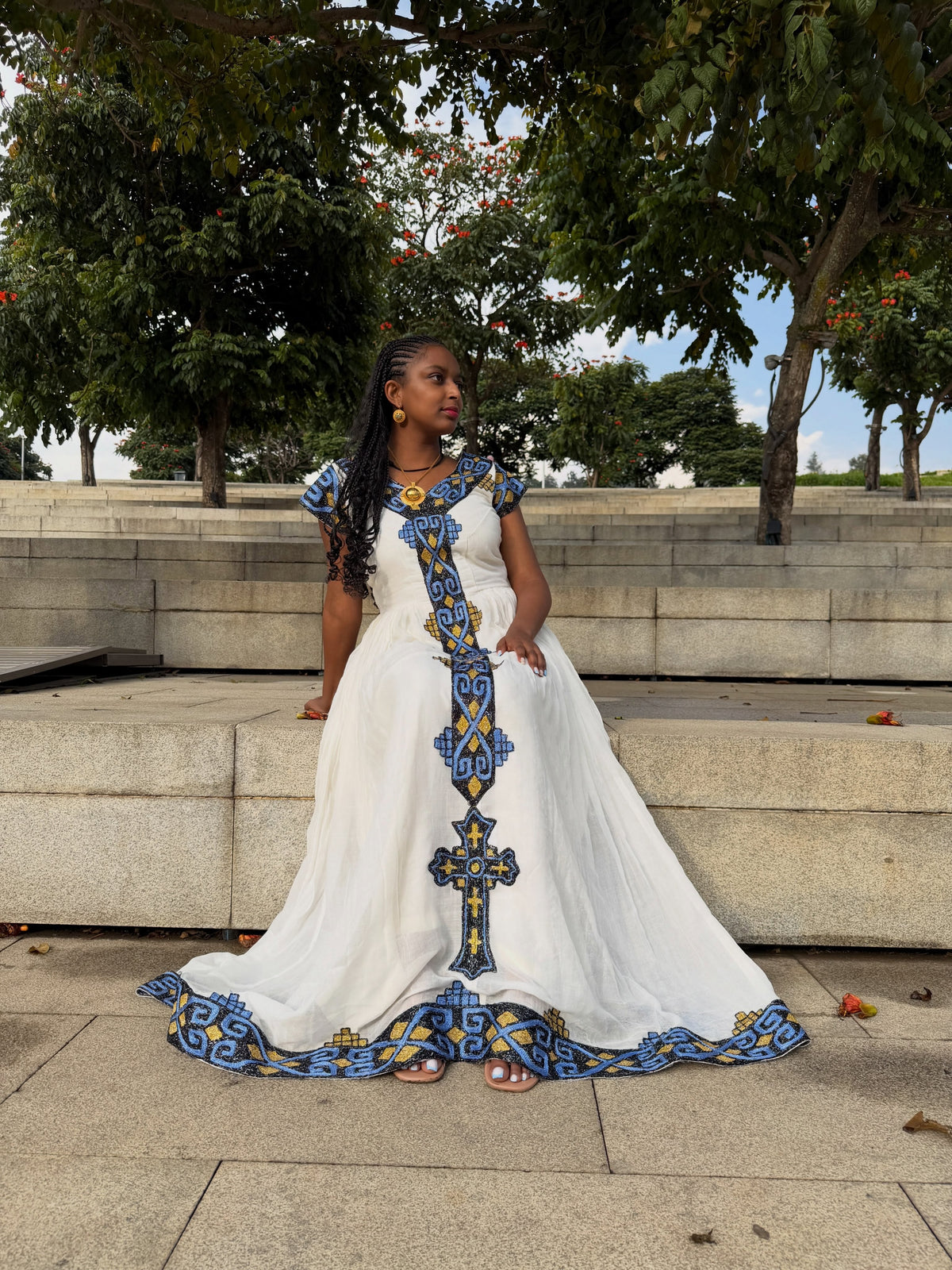 Woman standing outdoors in a white Ethiopian Dress featuring blue and gold woven embroidery down the front center and along the hem.