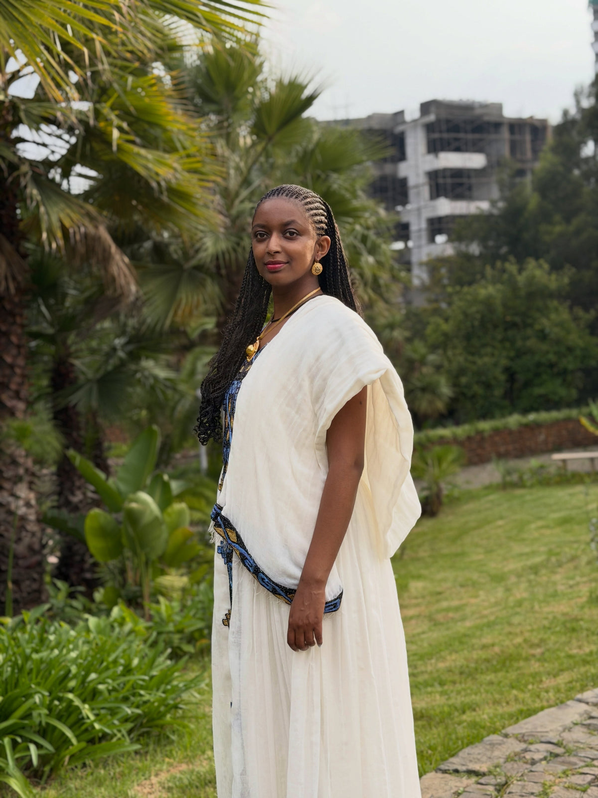 A woman wearing a white draped tunic Ethiopian Dress with a blue patterned sash, standing outdoors in a garden with palm trees and a building in the background.