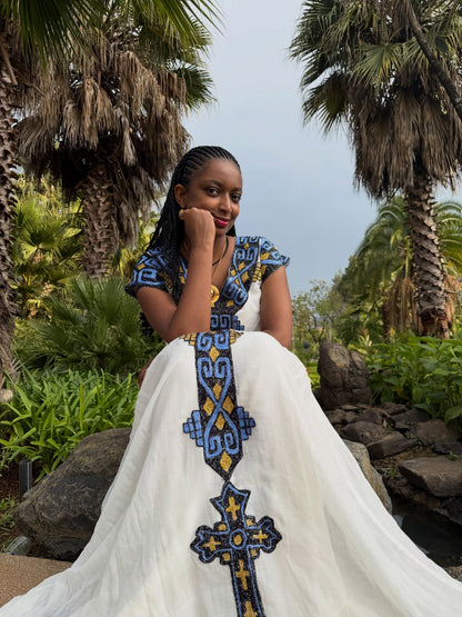 A woman wearing a white African  Ethiopian Dress with blue and yellow geometric patterns, posing outdoors in front of palm trees and rocks.