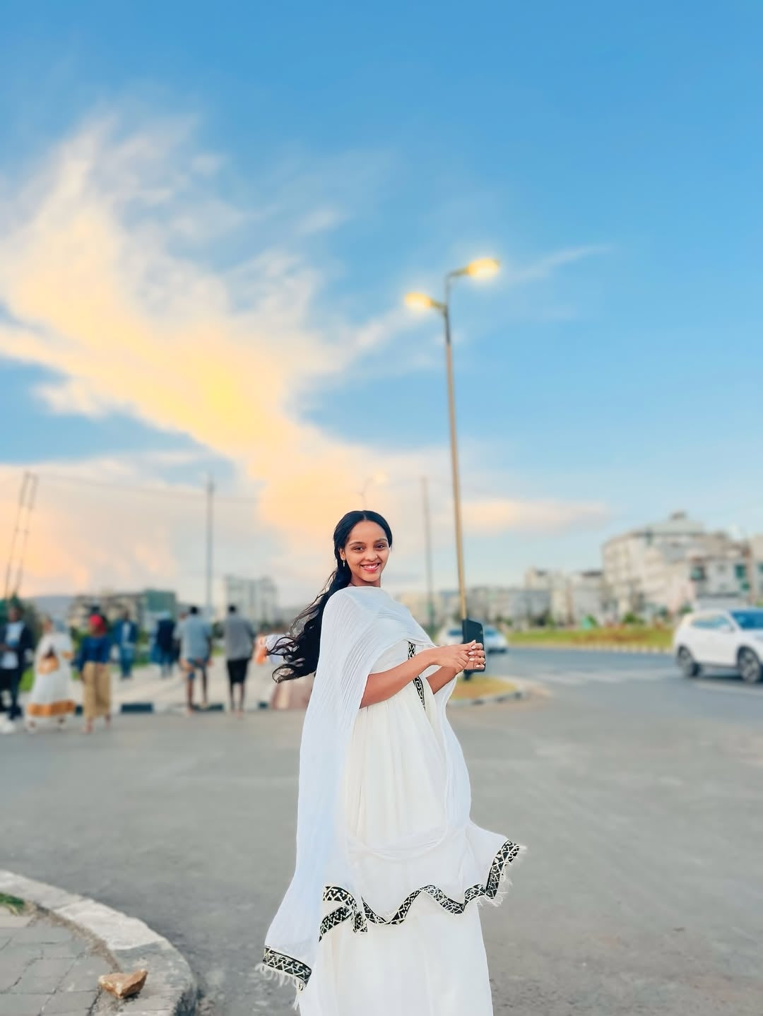 Woman in a white Traditional Dress standing on a street with a cityscape and sunset in the background