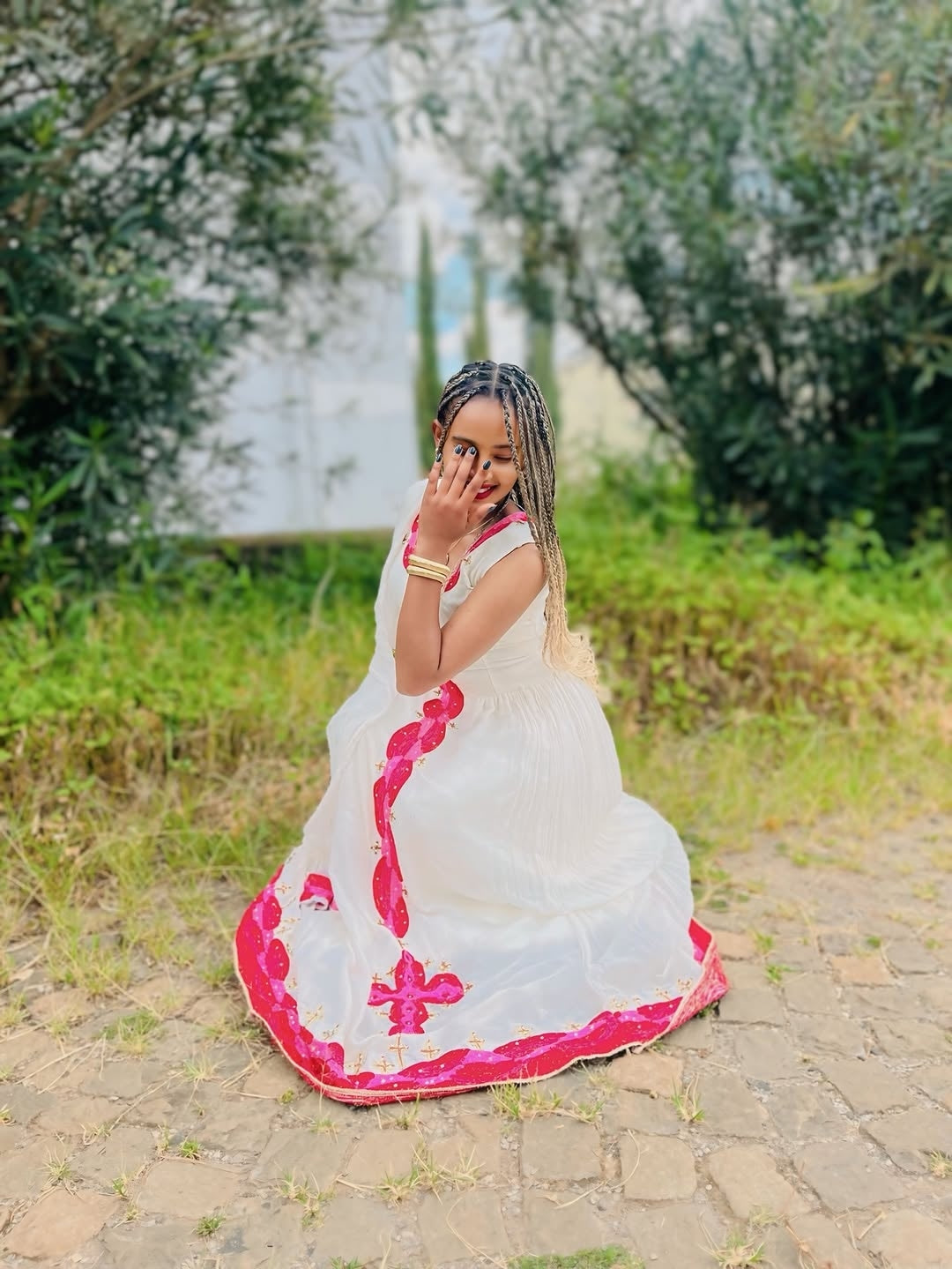 Woman in a white and pink traditional Habesha libs sitting outdoors with greenery in the background