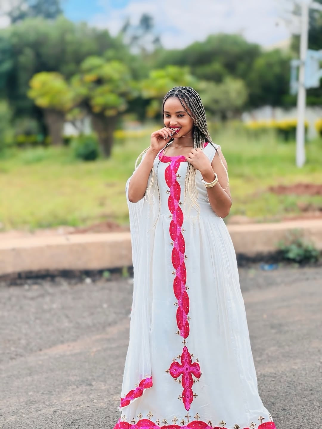 Woman in a white Habesha libs with pink embroidery standing outdoors.