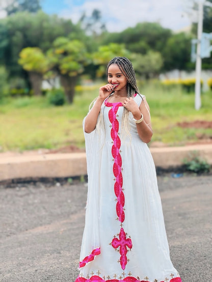 Woman in a white Habesha libs with pink embroidery standing outdoors.