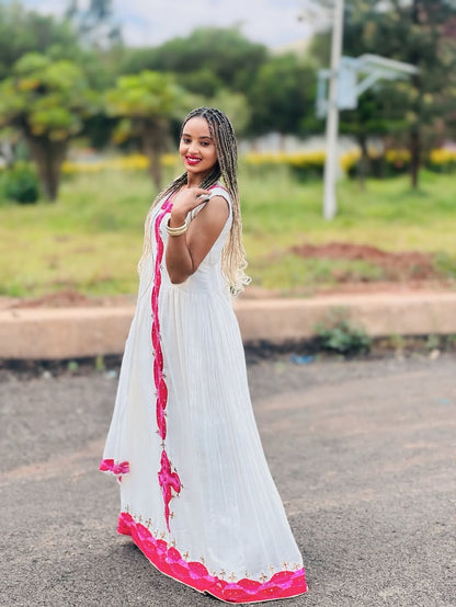 Woman in a white Habesha libs with pink accents standing outdoors on a road.