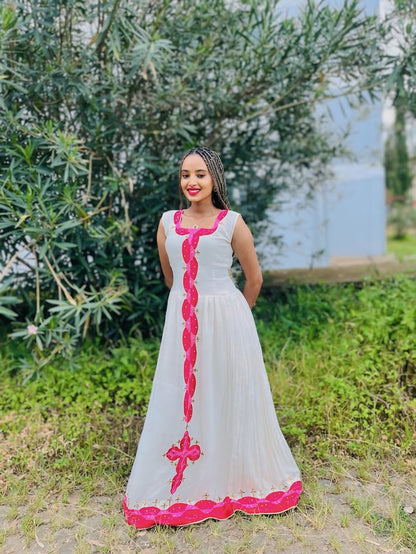 Woman in a white Habesha dress with pink accents standing outdoors near greenery and water.