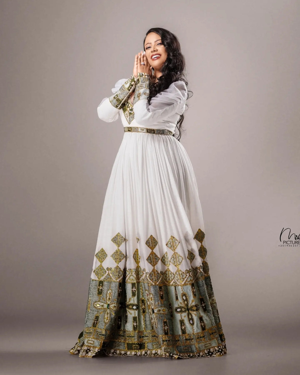 A woman modeling a long white traditional Habesha Wedding Dress  with ornate gold and green embroidery, standing against a plain taupe studio background.