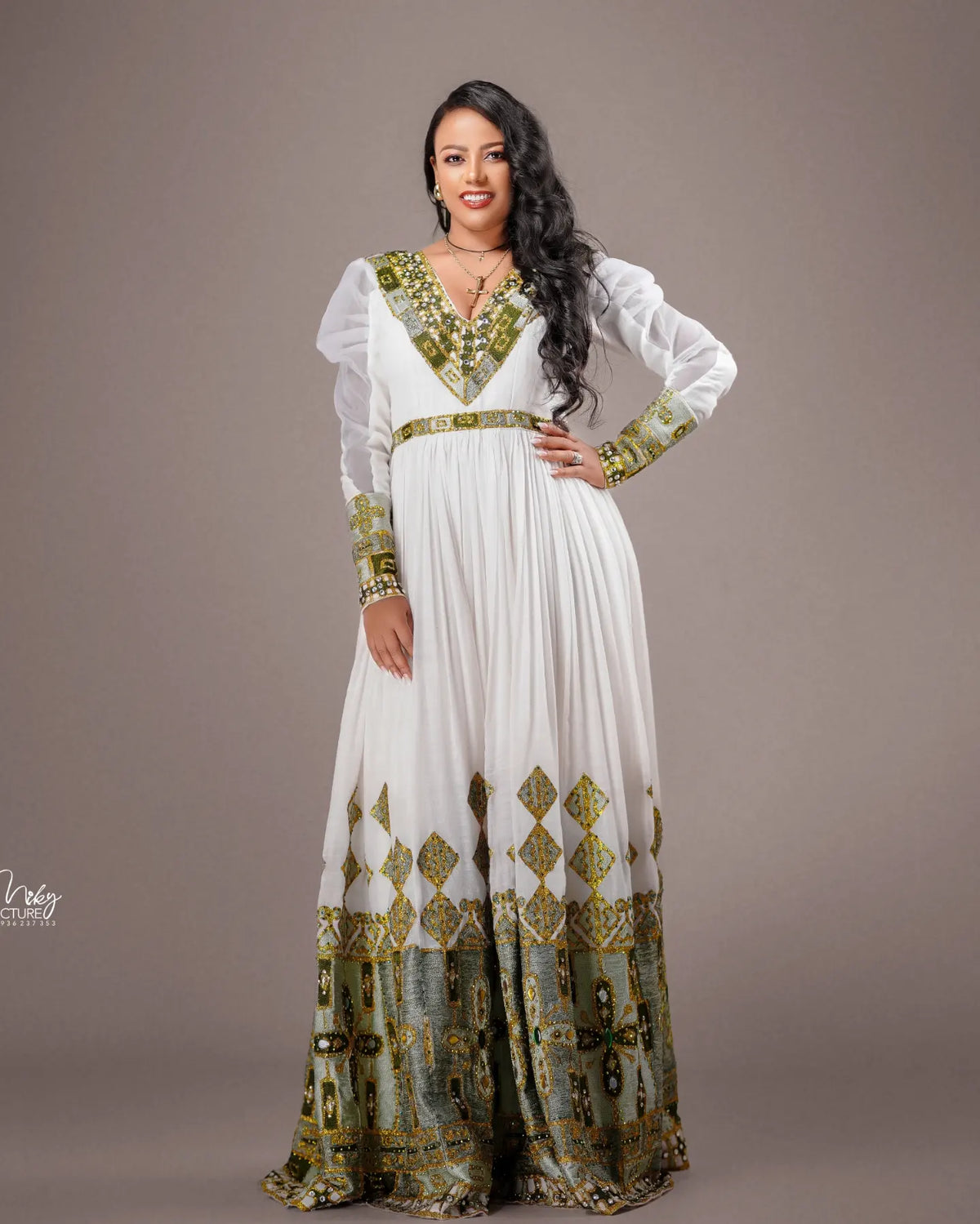 A woman modeling a long white Habesha Wedding Dress  with ornate green and gold embroidered patterns on the neckline, sleeves, waist, and hem, posed against a solid taupe studio background.
