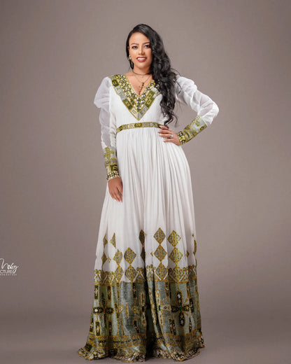 A woman modeling a long white Habesha Wedding Dress  with ornate green and gold embroidered patterns on the neckline, sleeves, waist, and hem, posed against a solid taupe studio background.