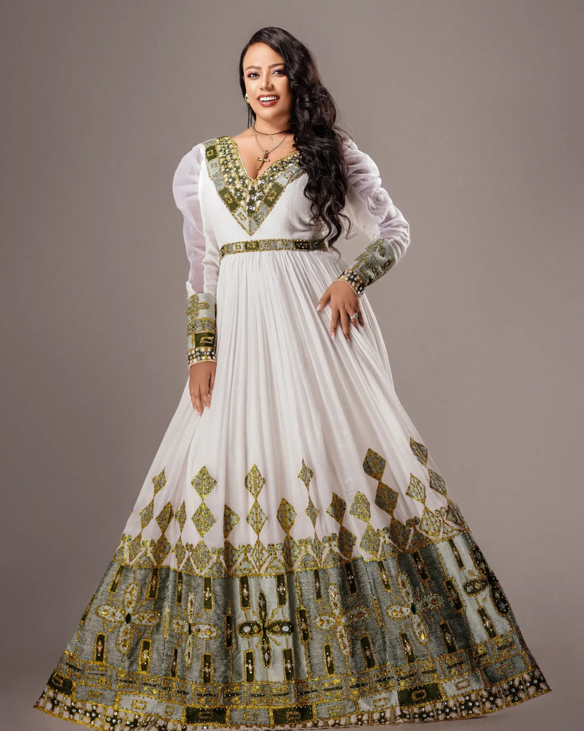 A woman modeling a long white traditional Habesha Wedding Dress  with extensive gold and green embroidery, featuring sheer long sleeves, a V-neckline, and a flared skirt with a highly detailed patterned hem.