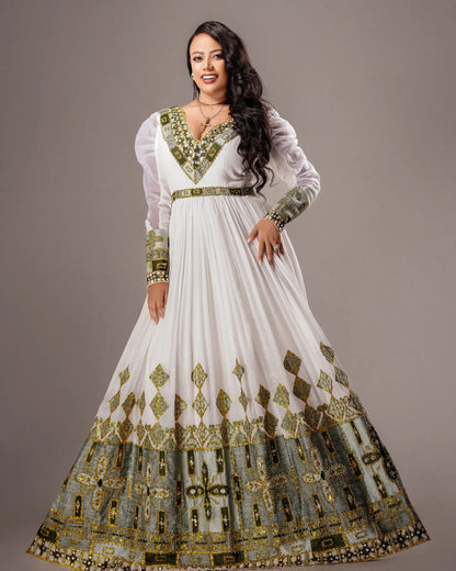 A woman modeling a long white traditional Habesha Wedding Dress  with extensive gold and green embroidery, featuring sheer long sleeves, a V-neckline, and a flared skirt with a highly detailed patterned hem.