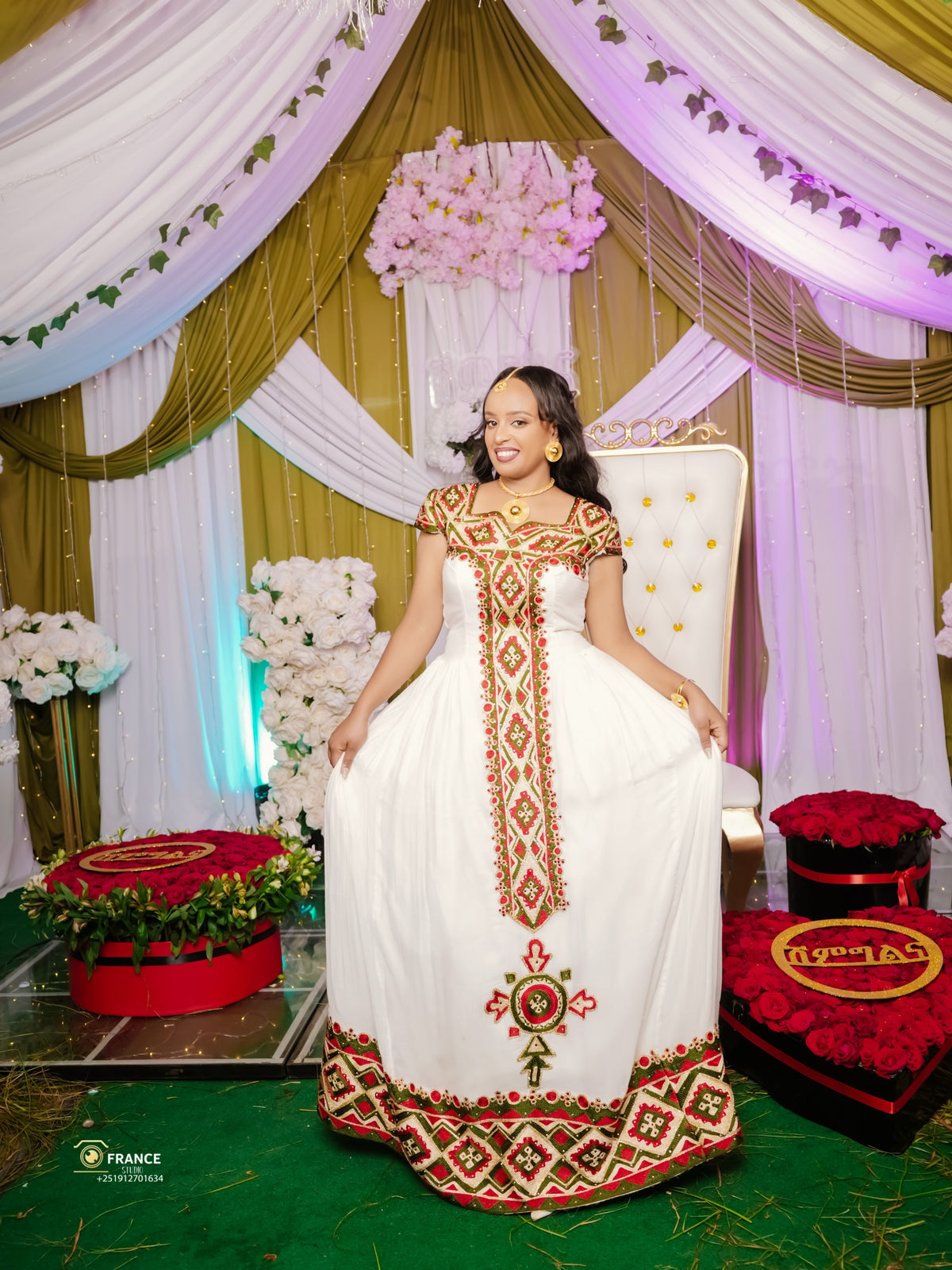 A woman wearing a traditional white  ethiopian dress with red and green geometric embroidery, standing in front of a decorated event backdrop with draped white and gold fabric, pink flowers, and a white chair.