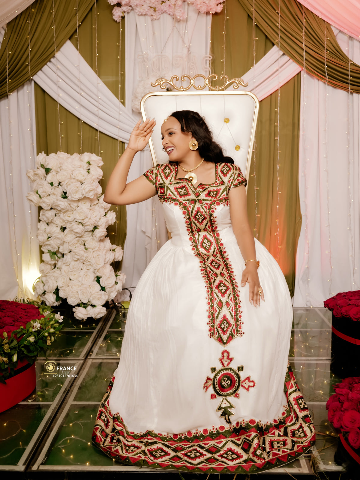 A woman wearing a white traditional ethiopian dress with red and green geometric embroidery poses on a white tufted throne chair in front of a decorated backdrop with gold drapes, white flowers, and red roses.