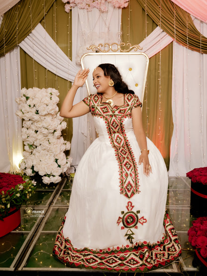 A woman wearing a white traditional ethiopian dress with red and green geometric embroidery poses on a white tufted throne chair in front of a decorated backdrop with gold drapes, white flowers, and red roses.