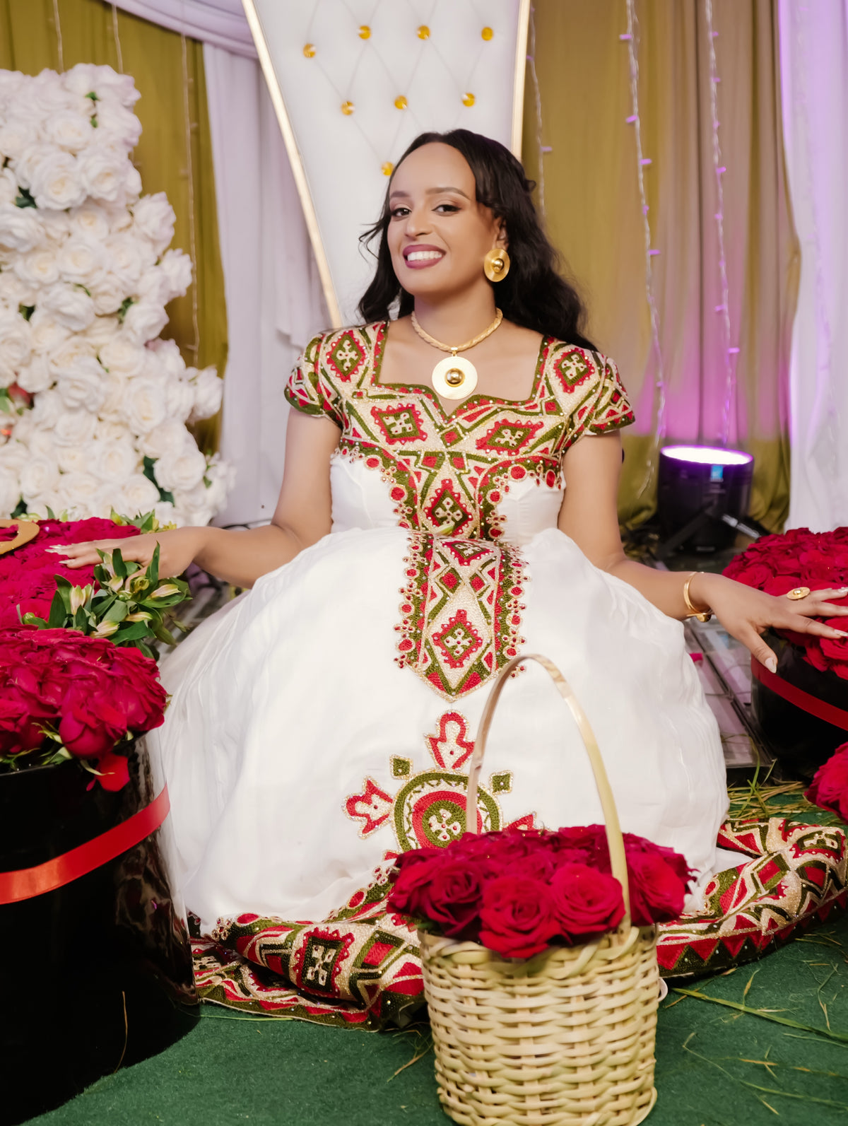 A woman wearing a traditional white ethiopian dress with red and green embroidered patterns sits in a decorated setting with red roses and a basket of roses in front of her.