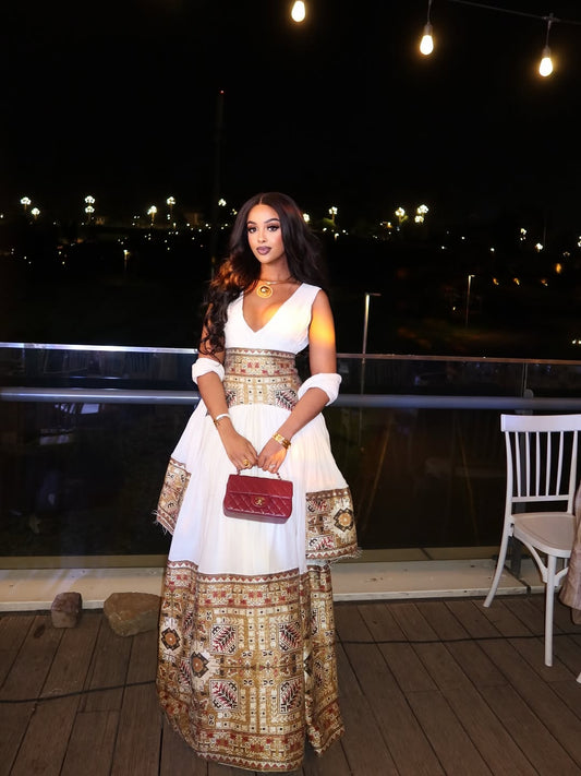 Woman in a long, patterned Ethiopia Kemis holding a red clutch on a wooden deck at night.
