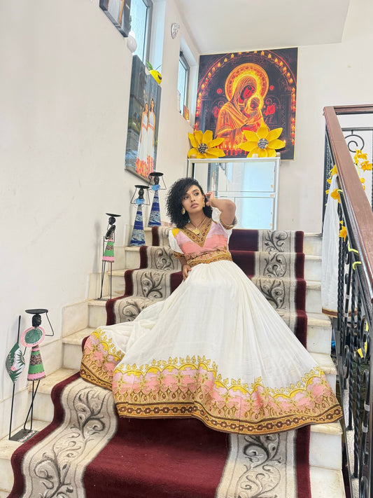 A woman wearing a traditional Indian salwar kameez Ethiopian National Dresses with a white skirt, pink blouse, and gold embroidery, posing on an indoor staircase decorated with patterned carpet runners and wall art.