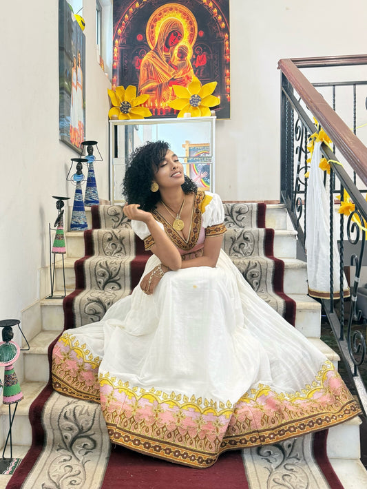 A woman wearing a white traditional Ethiopian National Dresses with gold and pink floral embroidery sits on a patterned staircase indoors. The dress has short sleeves, a V-neckline, and ornate gold detailing.