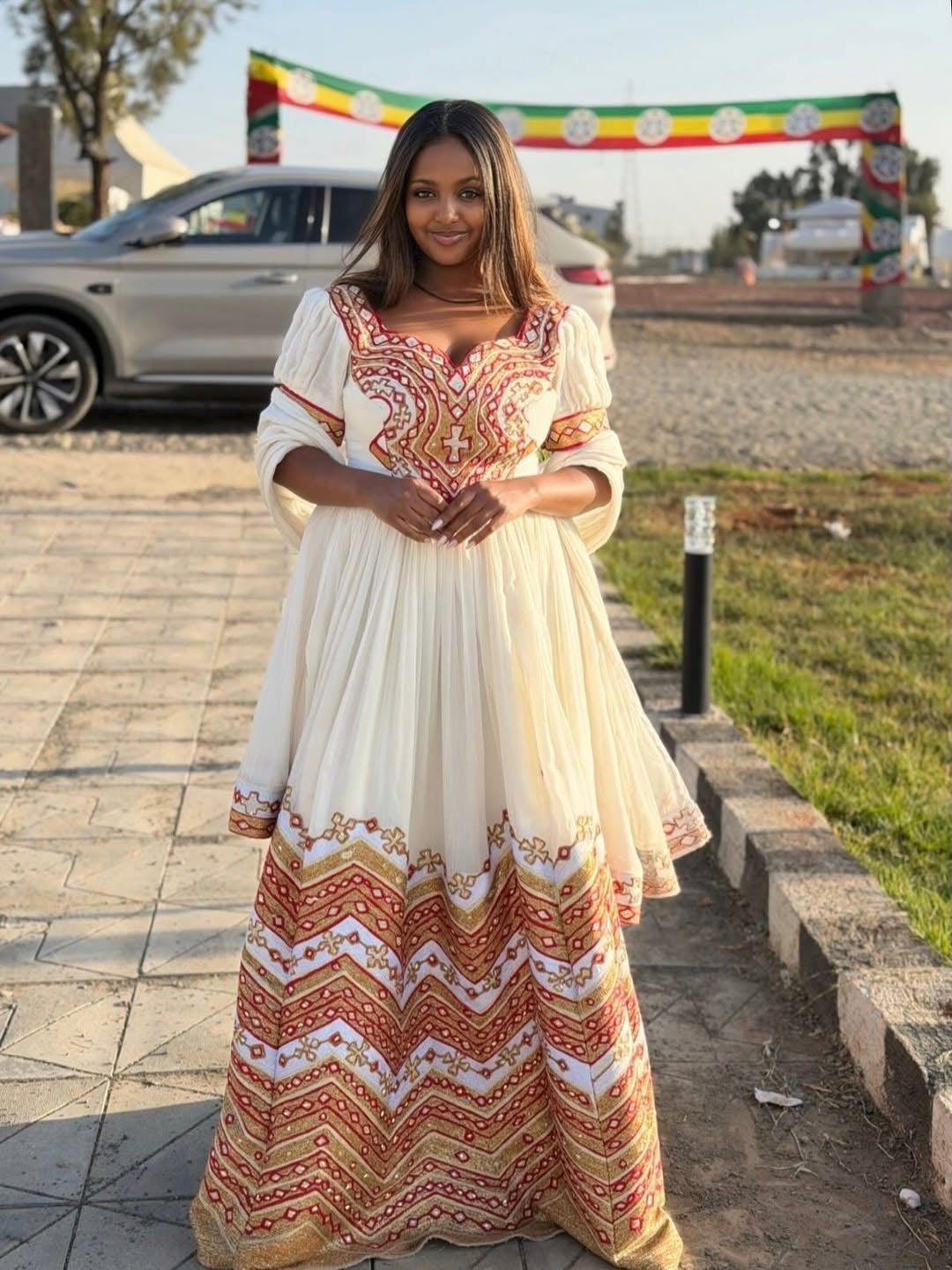 Woman in a long, patterned Eritrean Dressstanding outdoors with a car and colorful banner in the background.