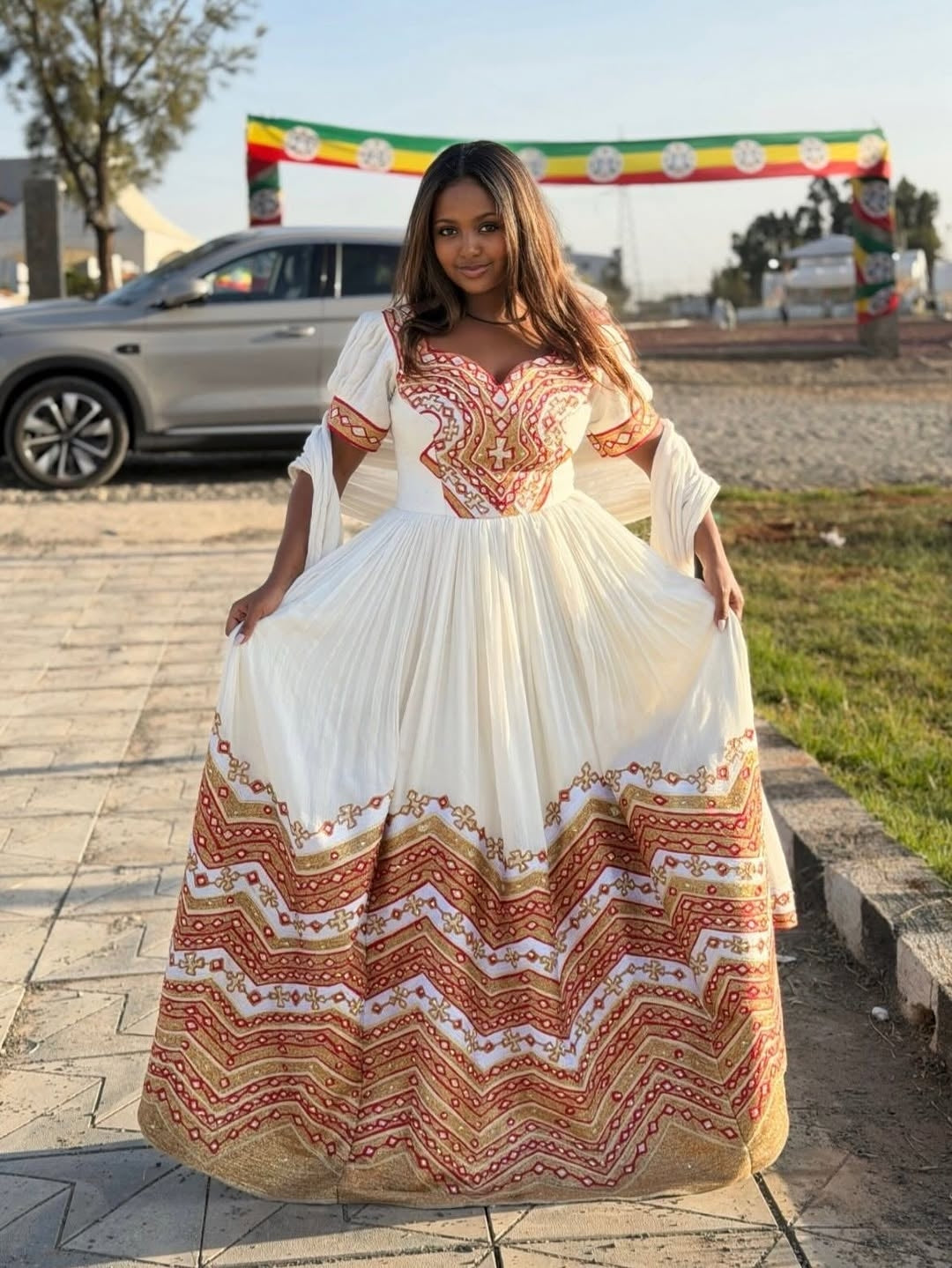 Woman wearing a long, patterned Eritrean Dress standing outdoors with a vehicle and banner in the background.
