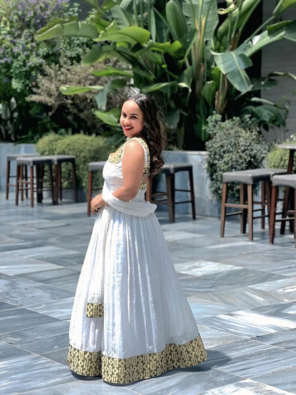 Woman in a white and gold traditional outfit standing outdoors with greenery in the background