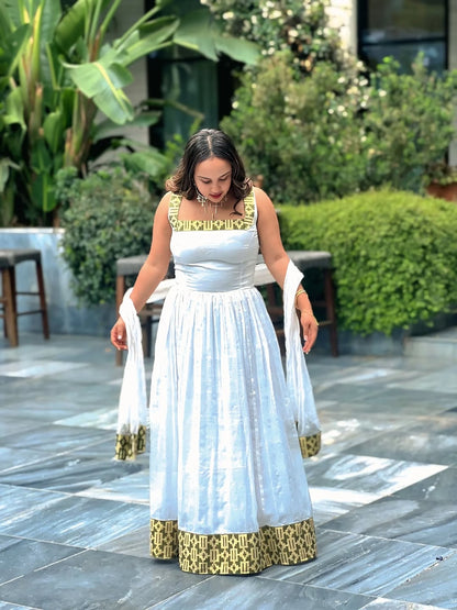 Woman in a white and gold traditional outfit standing outdoors with greenery in the background